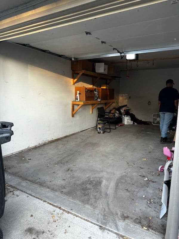 Empty garage with a cluttered corner, wall shelves, and a chair. Light overhead.