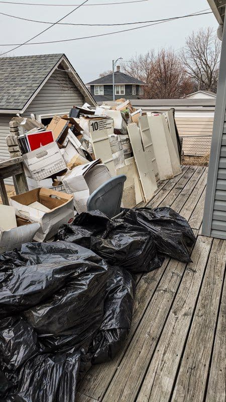 Piled cardboard boxes and black garbage bags on a wooden deck outside a house.